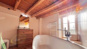 Et badeværelse med træloft, et stort badekar, et trækommode, et spejl og et vindue med sollys.