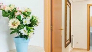 Un pasillo con una puerta que da a otra habitación, un cuadro en la pared y un jarrón azul con flores sobre una superficie blanca.