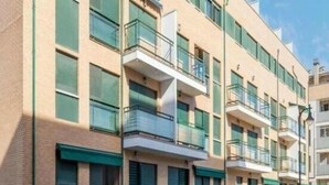 Un edificio residencial de varios pisos con balcones, un coche estacionado frente a él y una cerca verde.