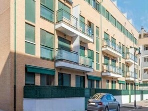 A multi-story residential building with balconies, a car parked in front, and a green fence.