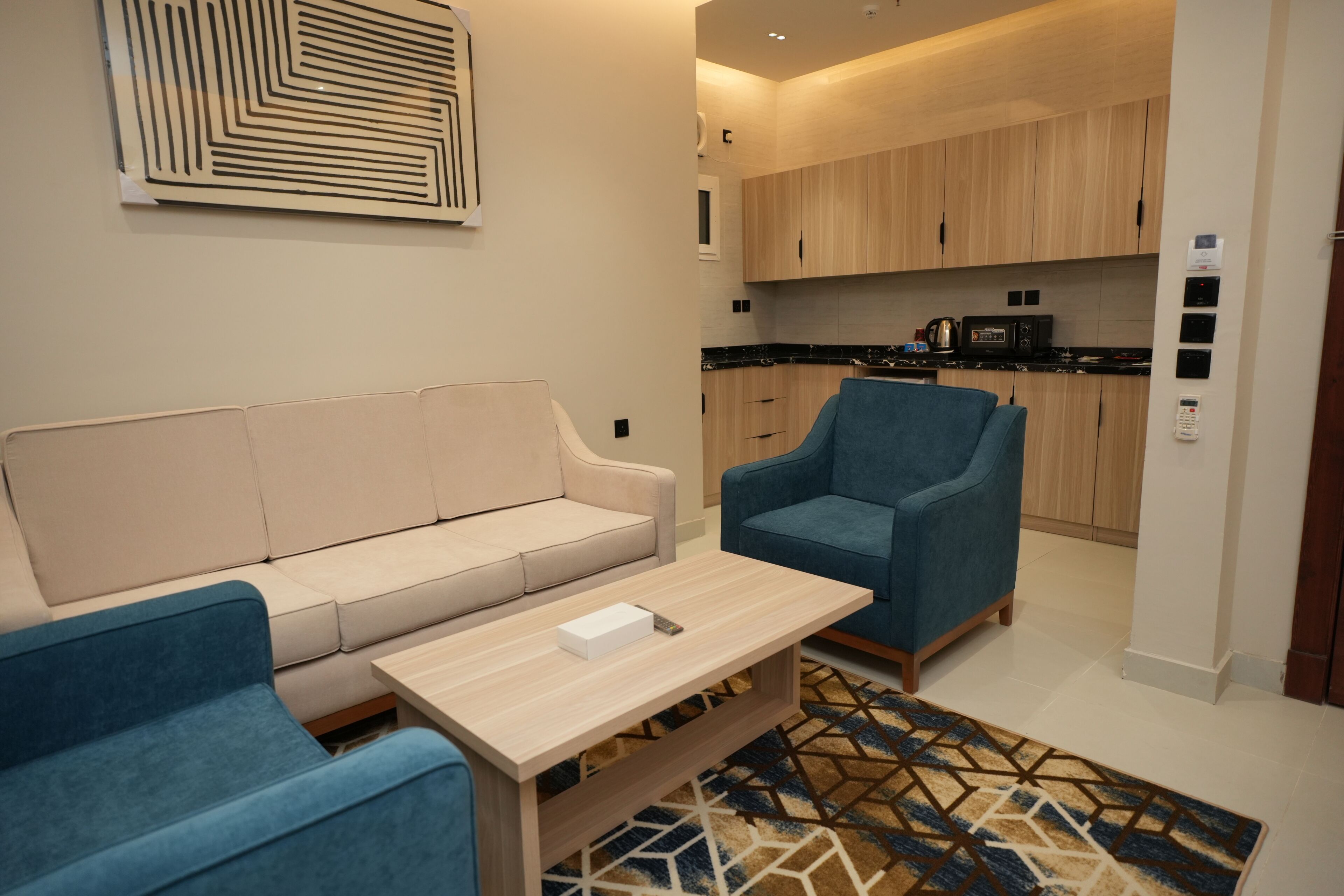 Un salon moderne avec un canapé beige, un fauteuil bleu et une table basse en bois. On aperçoit une cuisine à l’arrière-plan, avec des meubles en bois et un four à micro-ondes.
