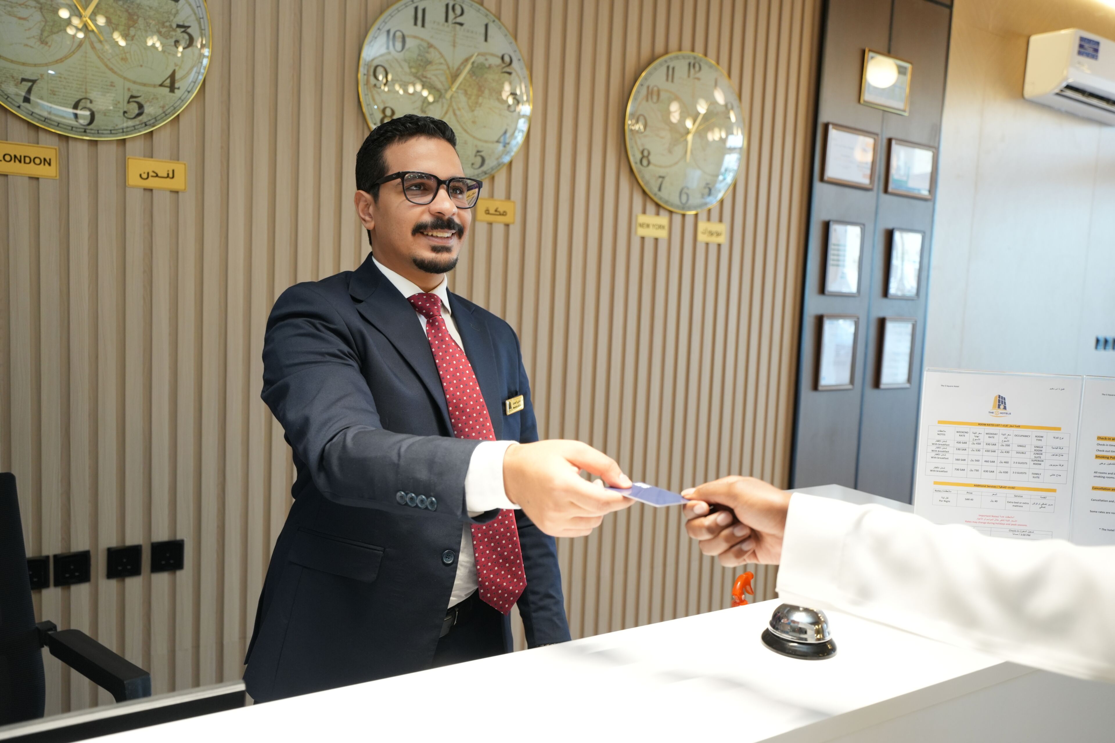 A hotel reception area with clocks on the wall, a reception desk, and a name tag on the man standing.