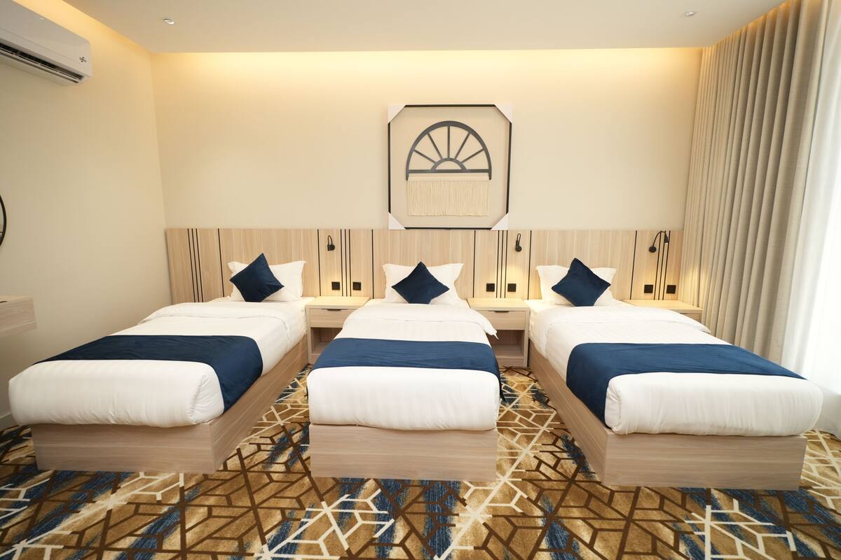 A hotel room with three single beds, a patterned carpet, a wall-mounted artwork, and a large window with curtains.