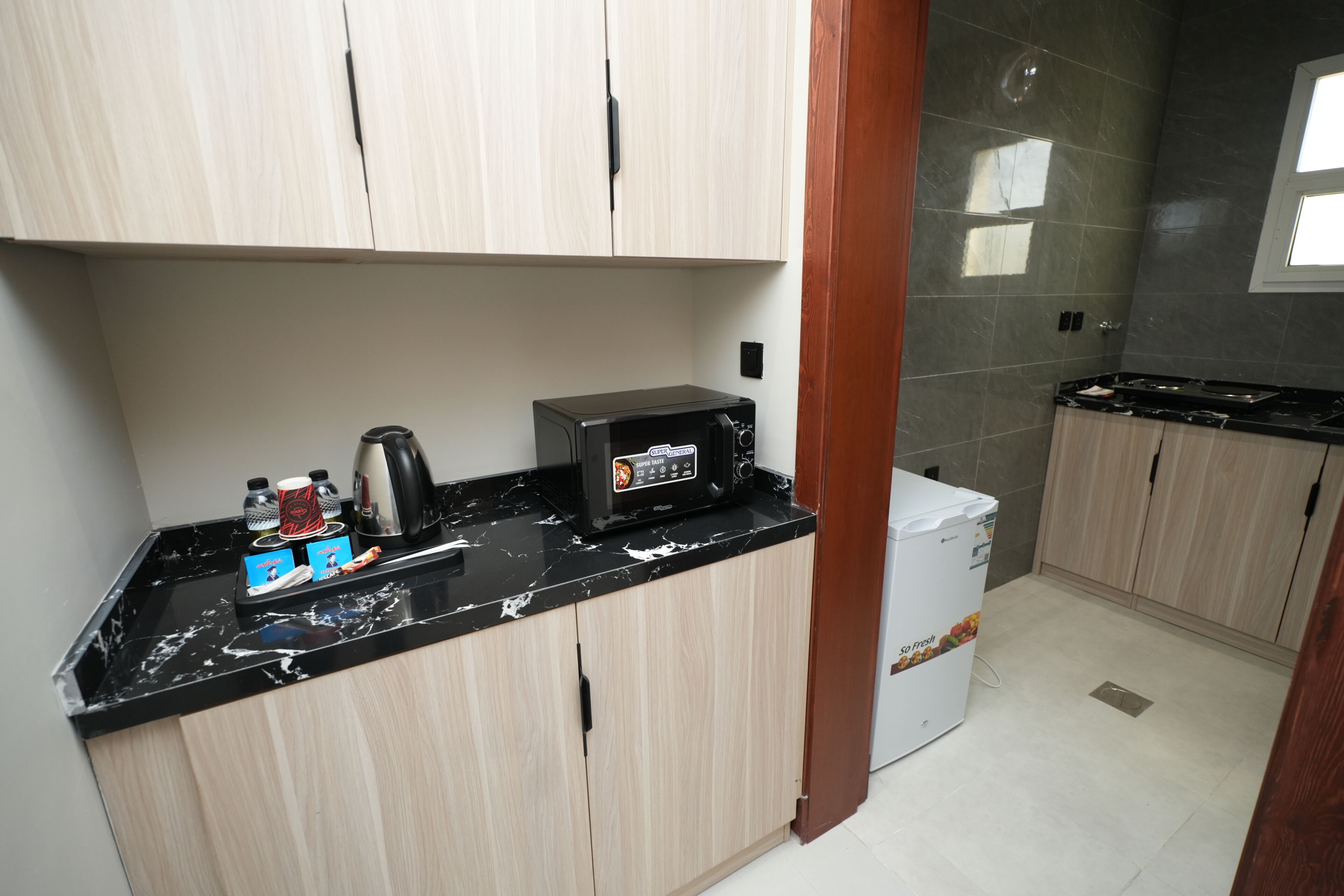 Uma cozinha compacta com bancada preta, micro-ondas, cafeteira e geladeira.