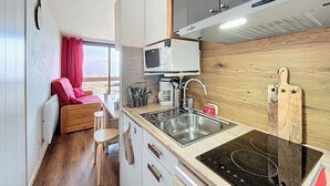 Ett kompakt kök med mikrovågsugn, diskho och spis. Det finns en matplats med ett träbord och en röd stol.