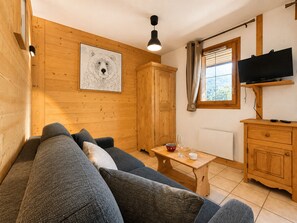 Una habitación acogedora con suelo de madera, un sofá, una mesa con vasos, un televisor y una ventana con cortinas.