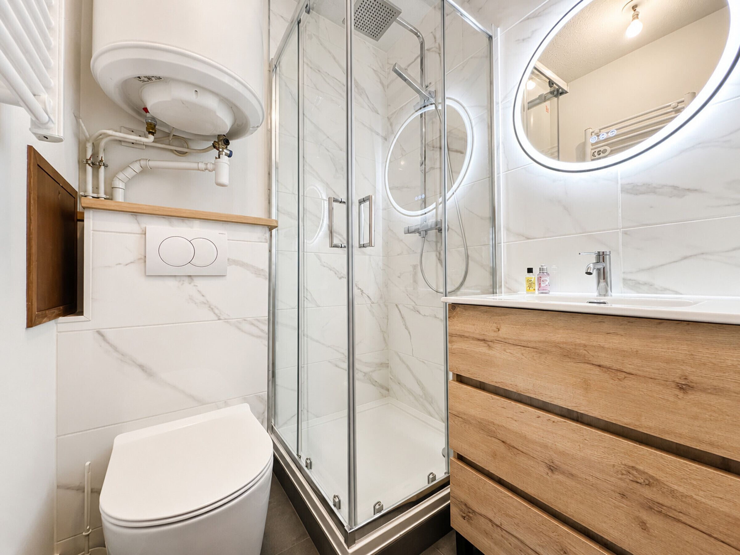 A modern bathroom with a glass-enclosed shower, a white toilet, a wooden vanity, and a round mirror.
