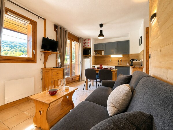 A cozy living area with a sofa, a wooden table, and a TV. There's a kitchen with cabinets and a window overlooking greenery.
