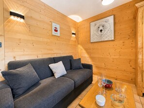 A cozy room with a grey sofa, wooden walls, a framed picture of a helicopter, and a table with glasses and a bowl. - Cozy studio cabin at ski slopes, 4 pers, balcony (Notre-Dame-de-Bellecombe)