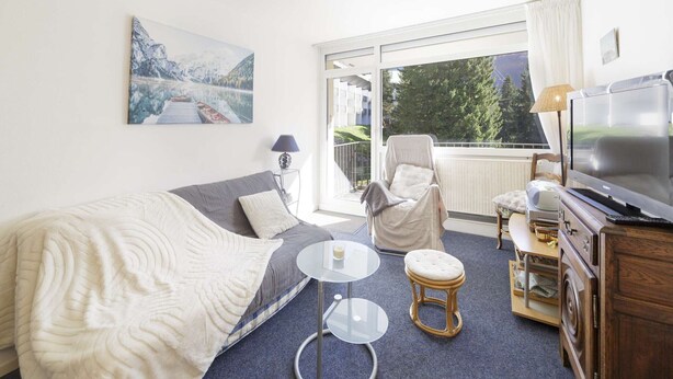 Et hotelværelse med seng, stol, lille bord, fjernsyn og et vindue med udsigt til grønne omgivelser.