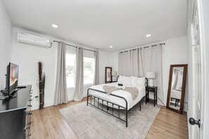 A modern bedroom with a bed, bedside tables, a TV, and a window with curtains. - The Lockwood, Residence One | EaDo by the Stadiums (Houston)