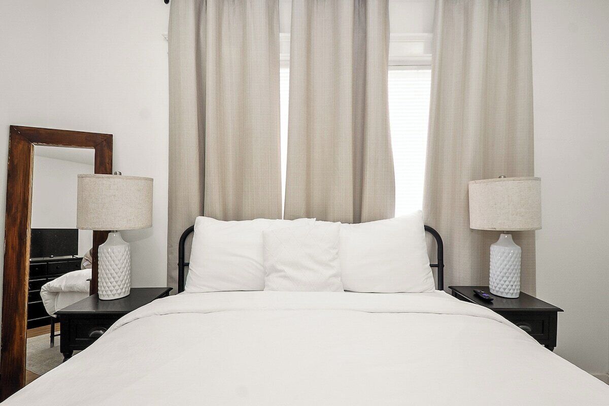 A hotel room with a bed, two bedside tables, a lamp on each table, a mirror on the left wall, and sheer curtains.