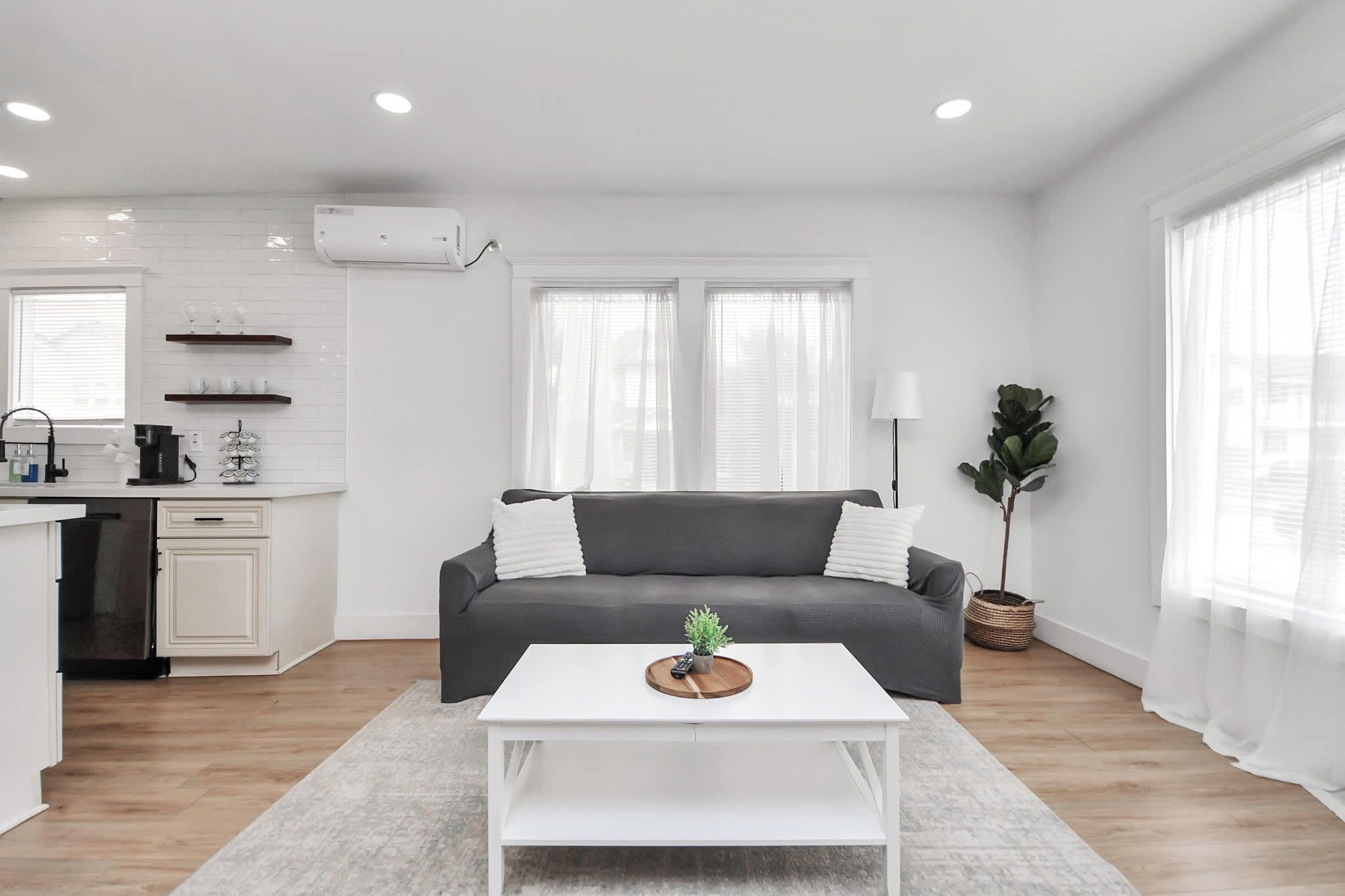 Un salon moderne avec un canapé gris, une table basse blanche et un coin cuisine doté de meubles blancs et d’un climatiseur intégré.