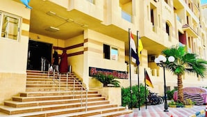 A hotel entrance with a staircase leading to the lobby, multiple flags, and a palm tree. - Florenze Khamsin Hurghada Room J113 - Room 9 (Hurghada)