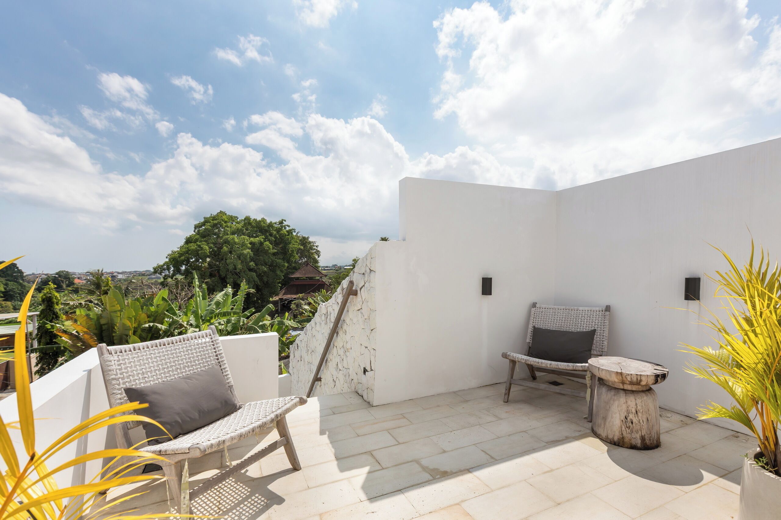 A rooftop patio with two wicker chairs and a small round table, surrounded by potted plants and a white wall.