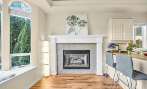 A kitchen with a fireplace, a dining area with chairs, and a window with a view of trees.