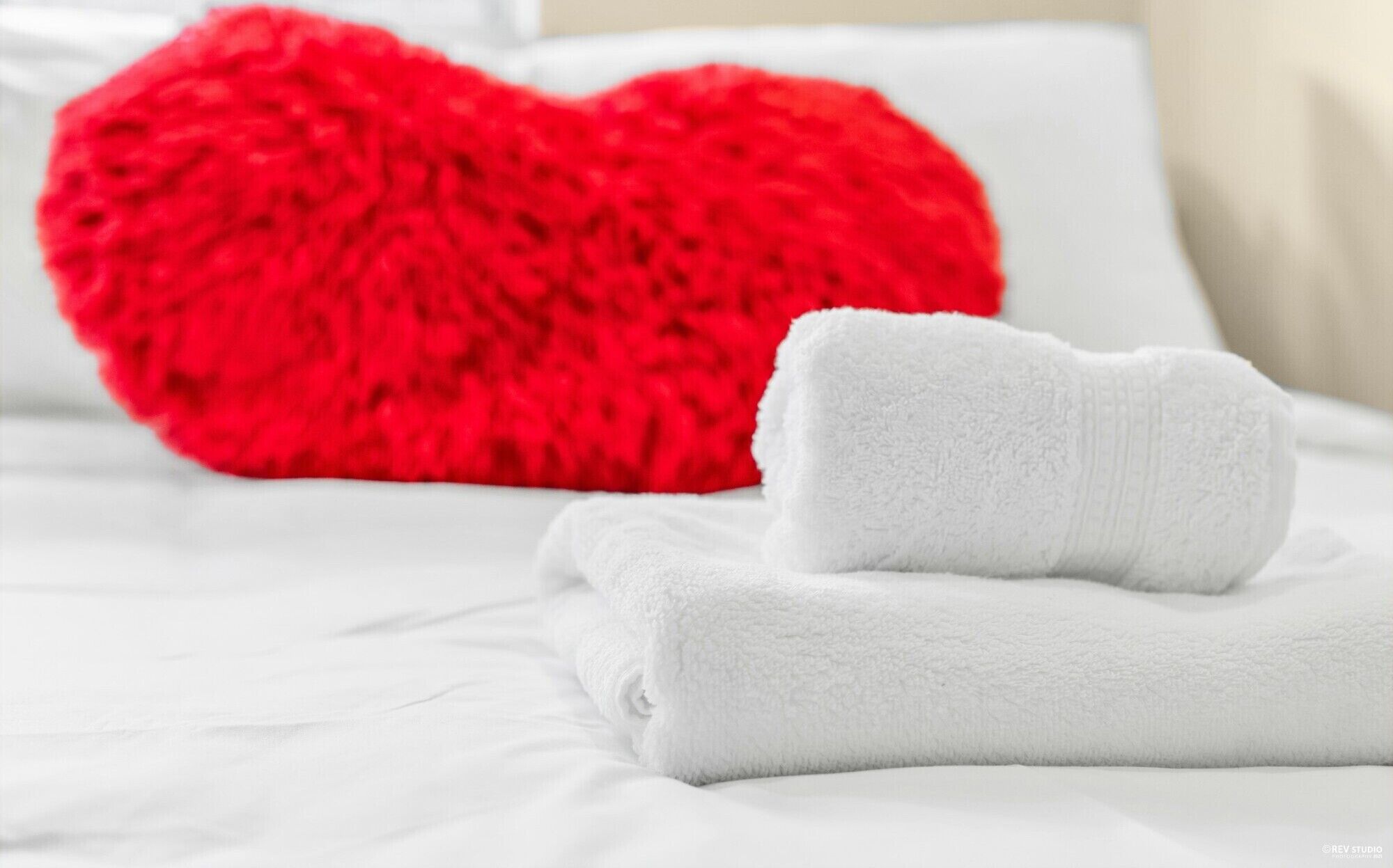 A neatly made bed with white linens and a red heart-shaped pillow.