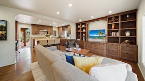 A spacious living area with a large sofa, a coffee table, and a bookshelf. There is a kitchen in the background with wooden cabinets and a dining area. - Majestic Corner Coastal Haven (San Diego)