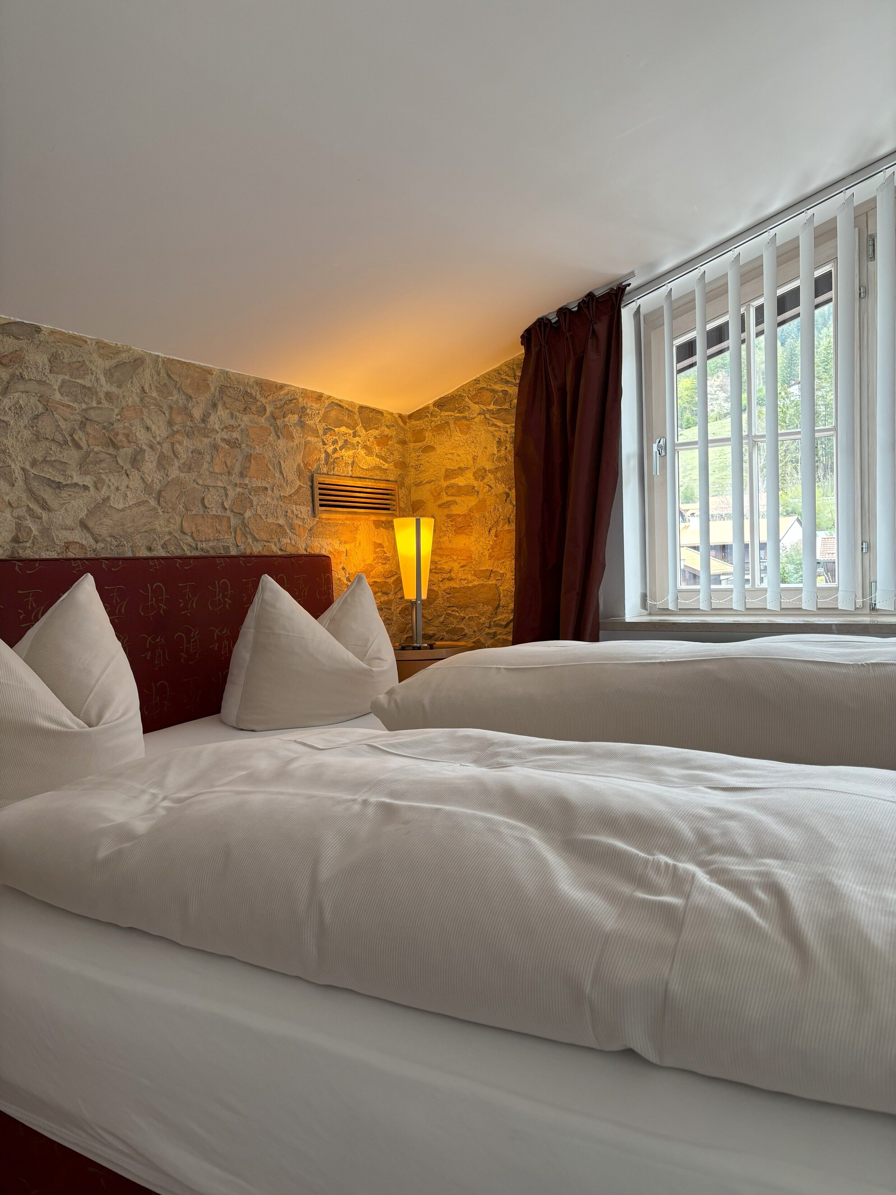 A hotel room with a stone wall, a bed with white bedding, a nightstand with a lamp, and a window with curtains.