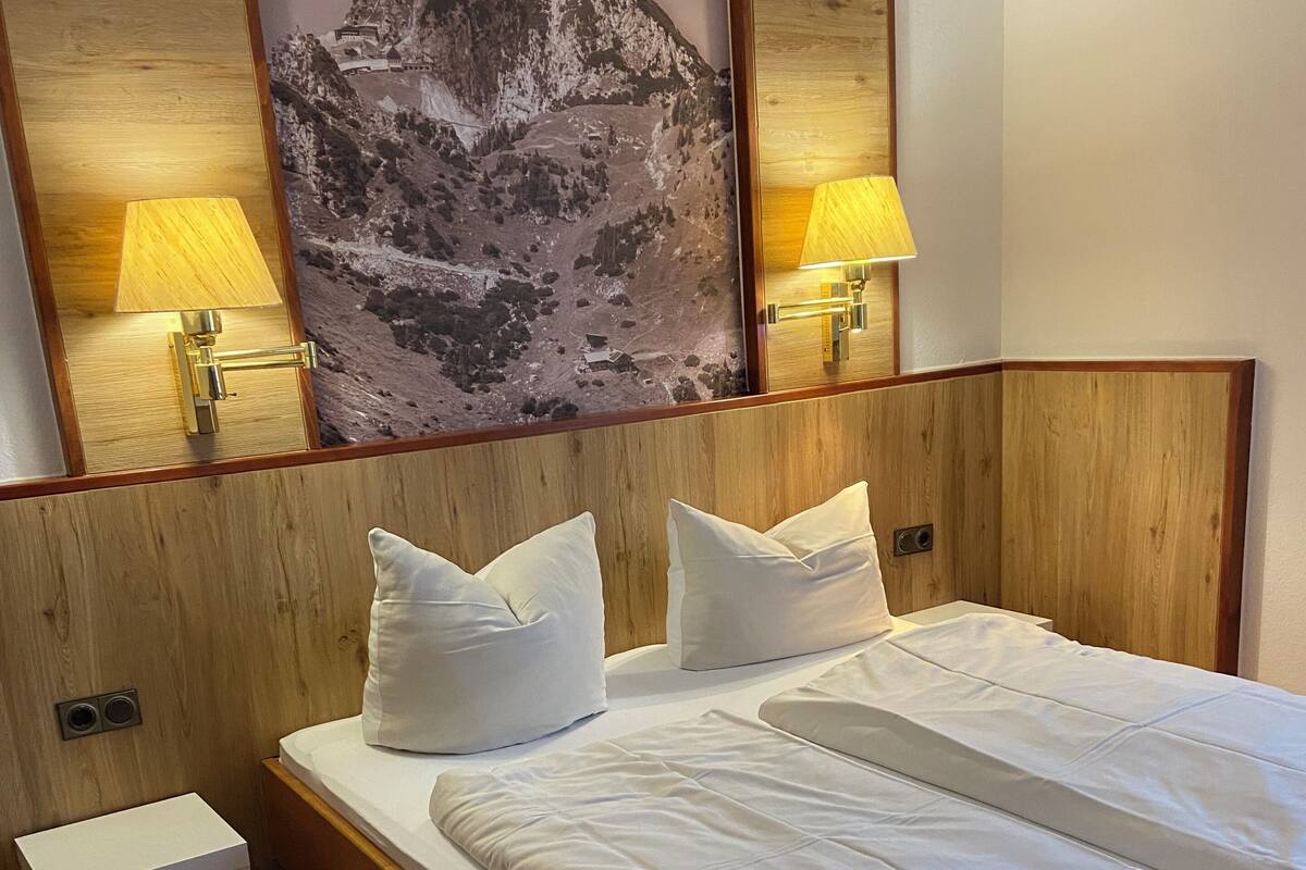 Een hotelkamer met een houten bed, witte beddengoed, twee bedlampen, een klein nachtkastje en een groot schilderij van een berg aan de muur.