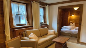 Een knus ingerichte kamer met een bank, fauteuil en een kleine tafel. In de achtergrond is een bed met hoofdbord te zien.