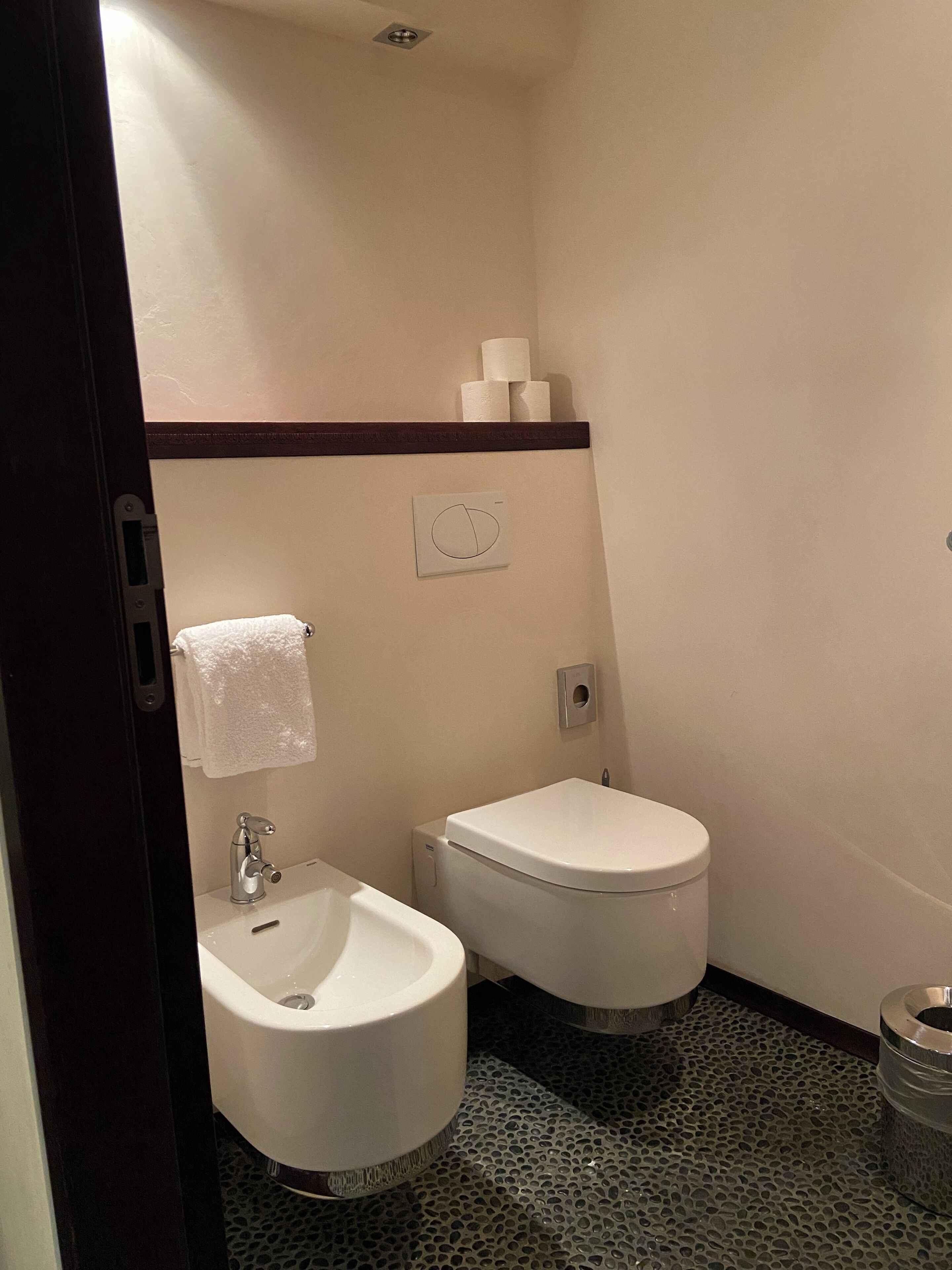 A small, compact bathroom with a sink, two toilets, and a towel rack.
