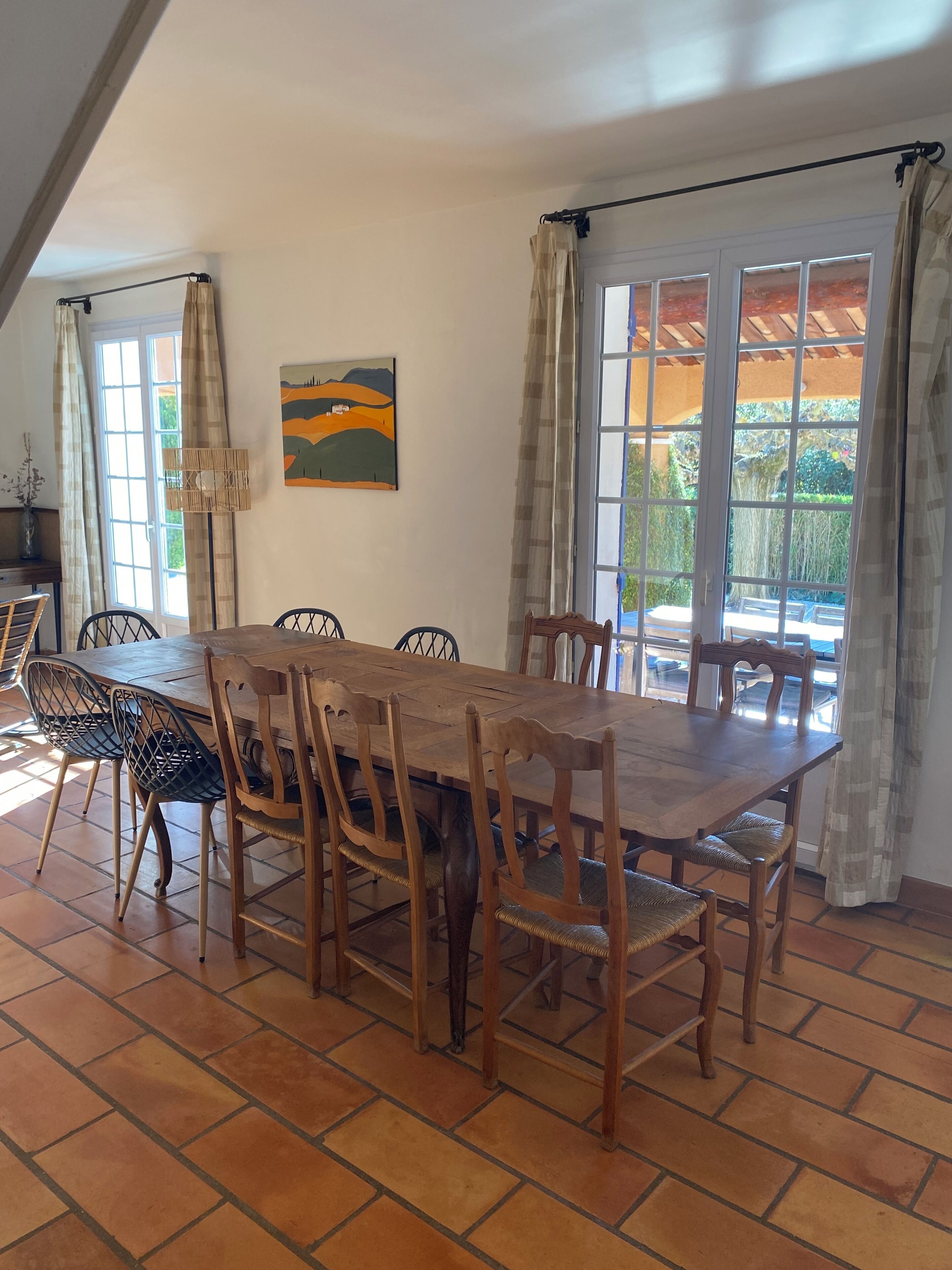 Un comedor con una mesa y sillas de madera, un cuadro en la pared y vistas a través de amplias ventanas.