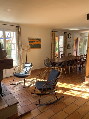 A dining area with a table and chairs, a rocking chair, and a view of the outdoors through large windows. - La Bambouseraie - Heated pool and nature (Lagnes)