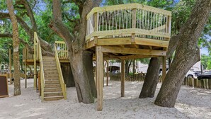 Una estructura de casa en árbol de madera con una escalera y una plataforma, rodeada de árboles y suelo arenoso.
