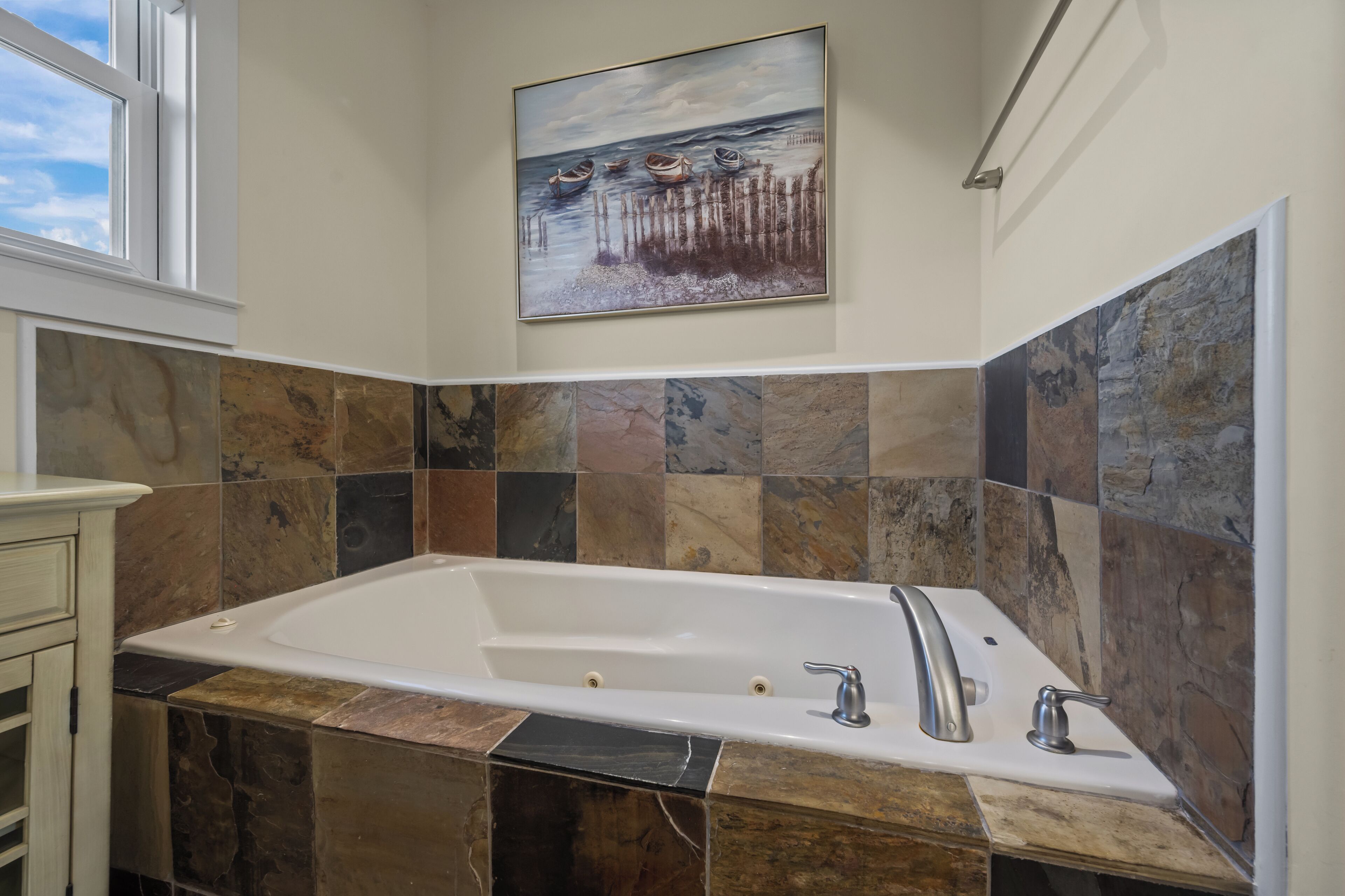 Un bagno con una vasca da bagno ampia, piastrelle in pietra e un quadro raffigurante barche al muro.