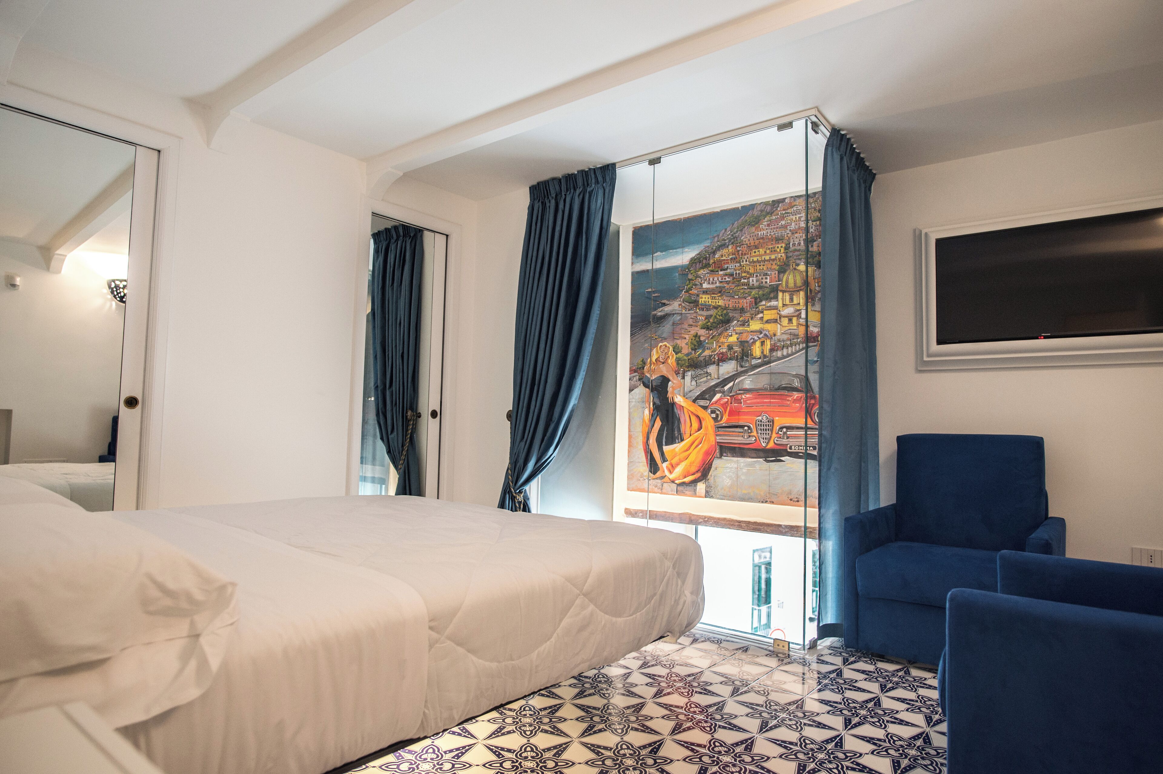 Una camera d'albergo con un letto, una poltrona blu, un quadro raffigurante una veduta costiera e un pavimento piastrellato.