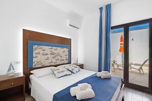 Une chambre d’hôtel avec un lit, une tête de lit ornée d’un motif sur le thème de la marine, un petit bureau et une vue sur l’espace extérieur aménagé en coin salon.