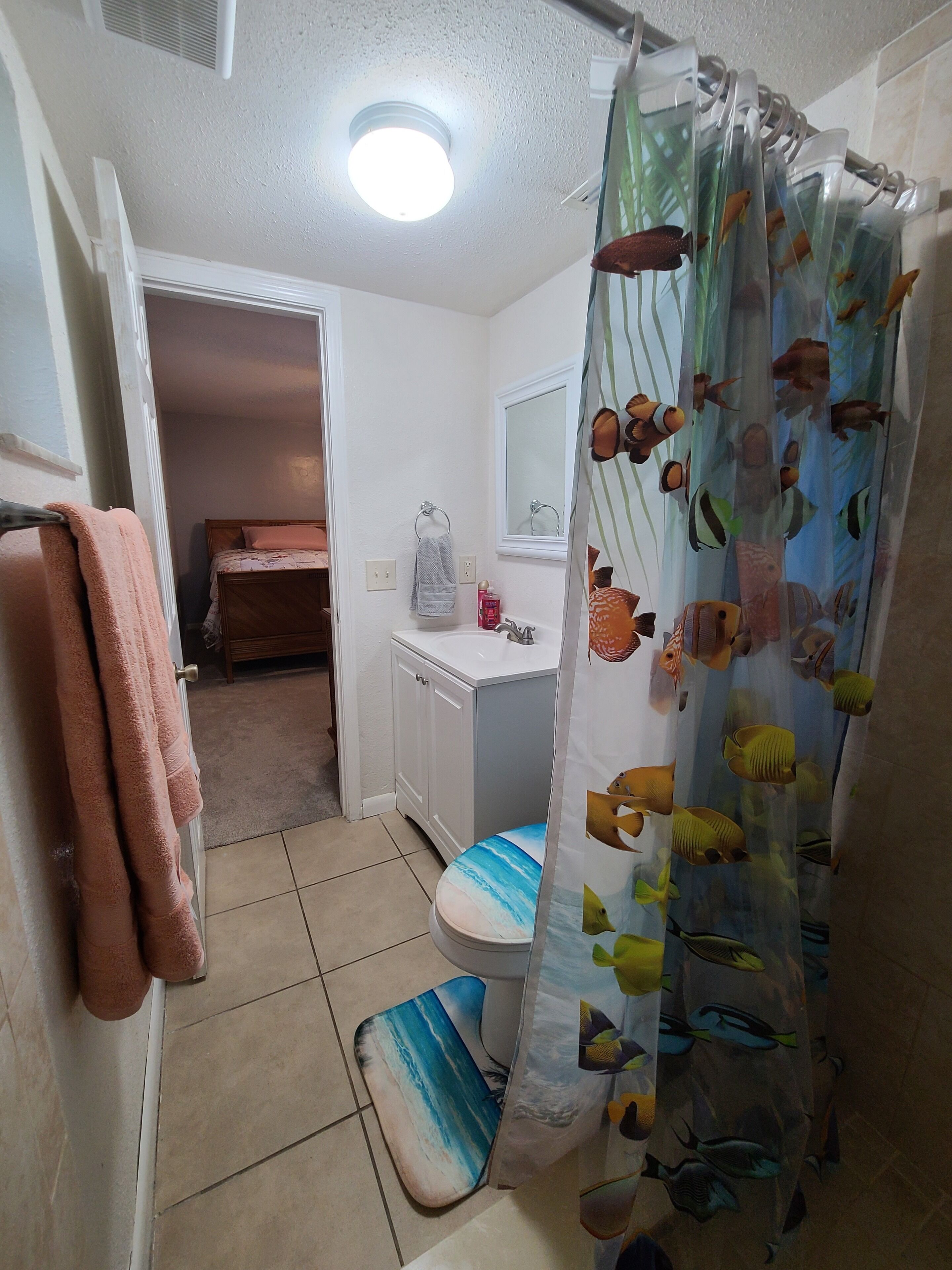 Ein Badezimmer mit Duschvorhang mit Fischmotiven, einer Toilette mit Sitzbezug im Strandstil, einem Waschbecken und einem Spiegel darüber.