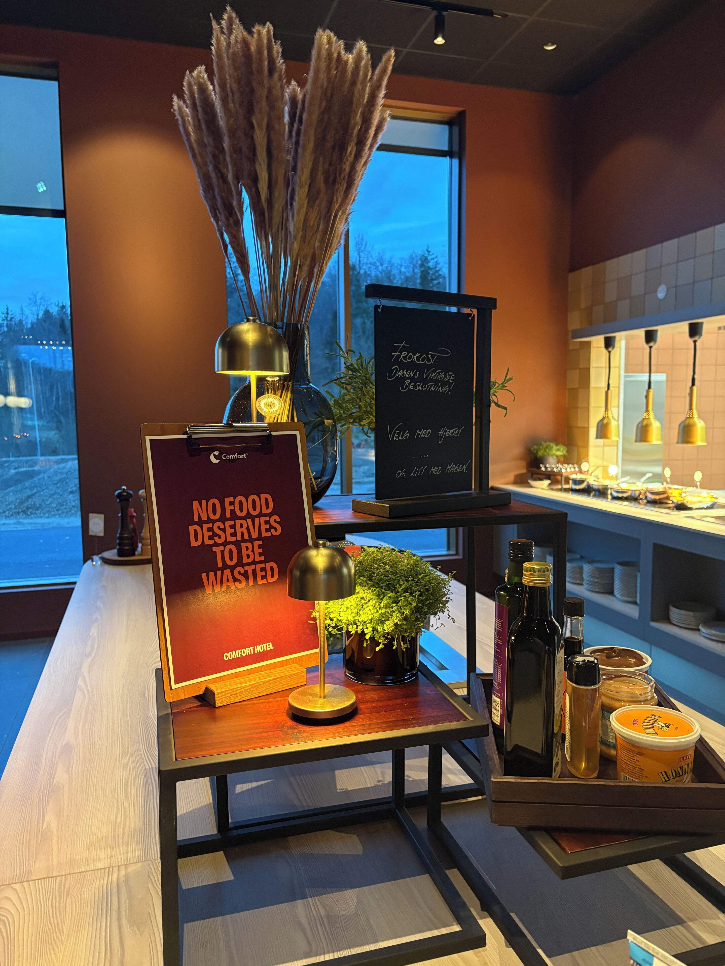 Zona de recepción de hotel con un cartel que dice 'NO SE DESPERDICIA NADA BUENO', un jarrón con plantas secas, una pizarra con texto y una barra con varios objetos.