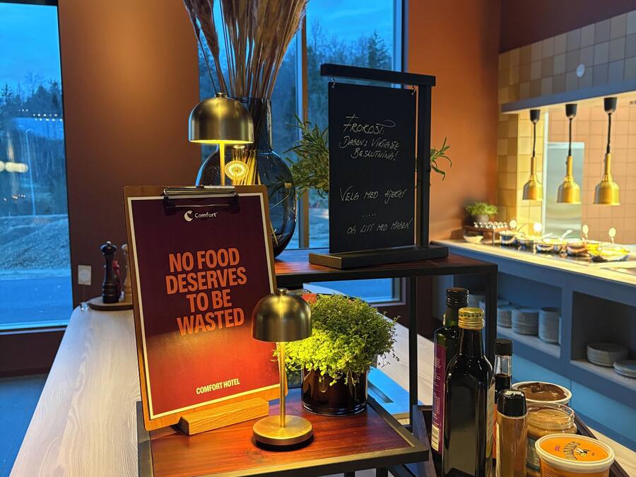 Zona de recepción de hotel con un cartel que dice 'NO SE DESPERDICIA NADA BUENO', un jarrón con plantas secas, una pizarra con texto y una barra con varios objetos.