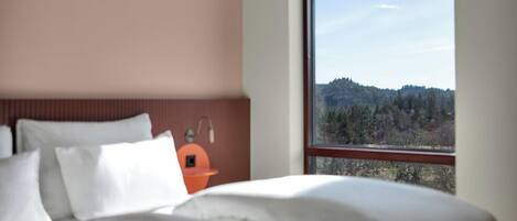 A bedroom with a large window, a bed with white bedding, a wooden headboard, and a small orange table with a lamp.