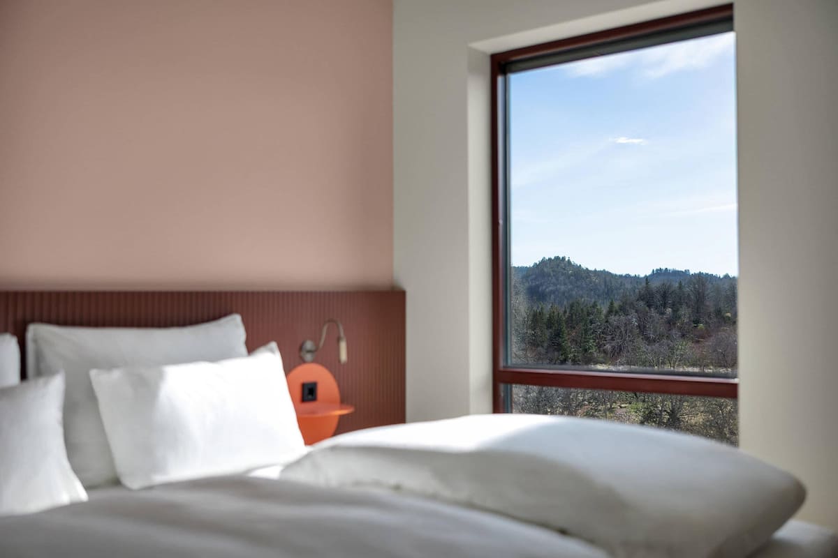 Ein Schlafzimmer mit Bett, Kissen, einem Nachttisch, einer Lampe und einem Fenster mit Blick auf Bäume und Hügel.