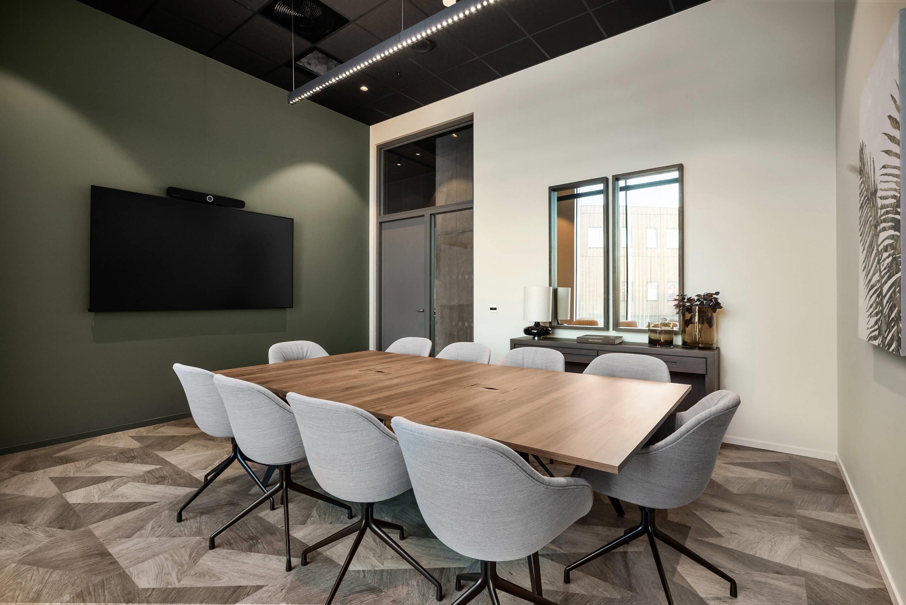 Una sala de conferencias con una amplia mesa de madera, seis sillas grises, un televisor de pantalla plana, un ventanal y una planta sobre un escritorio.