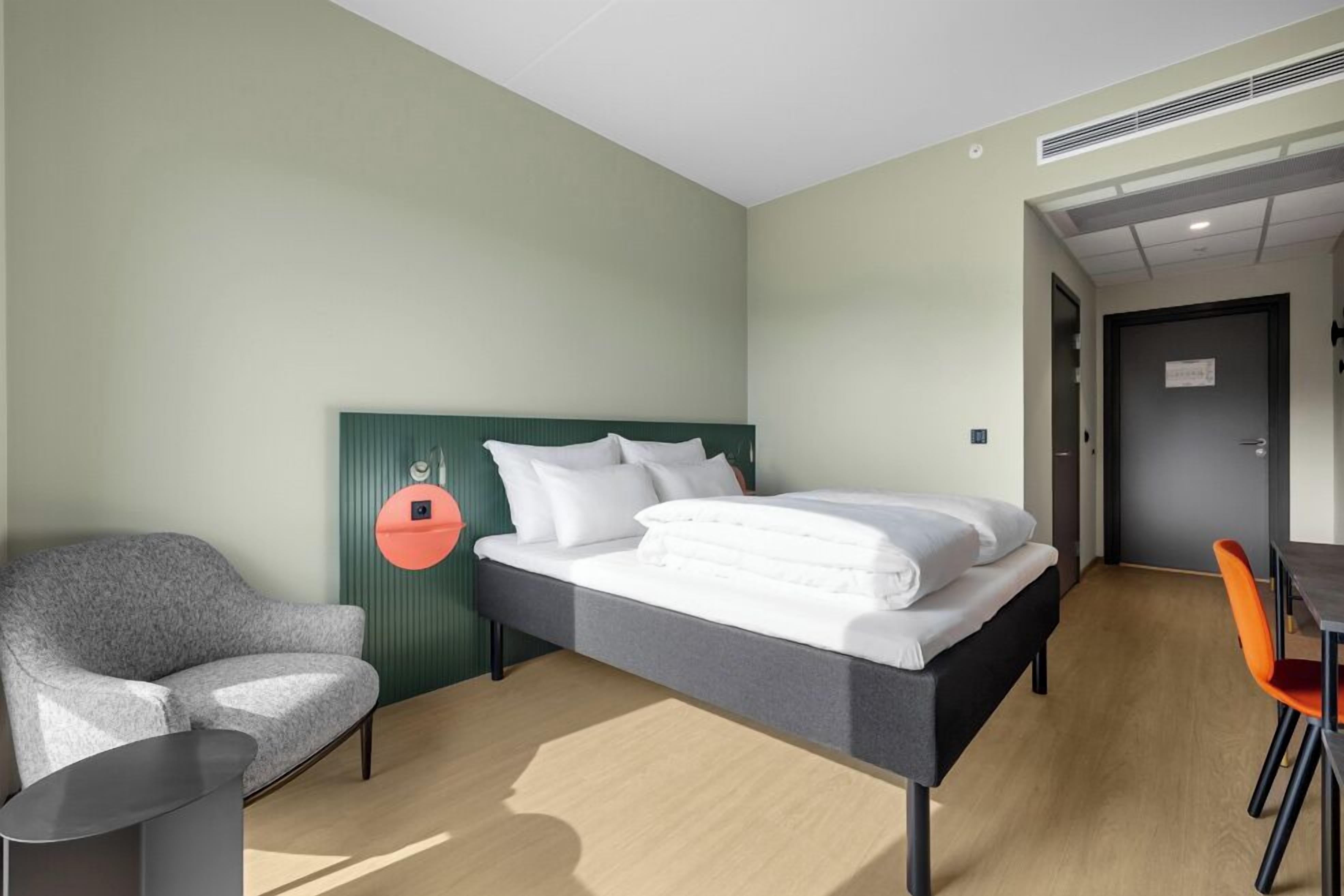 A hotel room with a bed, a chair, a table, and a door.