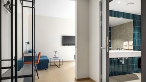 A modern hotel room with a glass partition, a blue sofa, a small table with a plant, a flat-screen TV, and a bathroom with a blue tiled wall and white sink.
