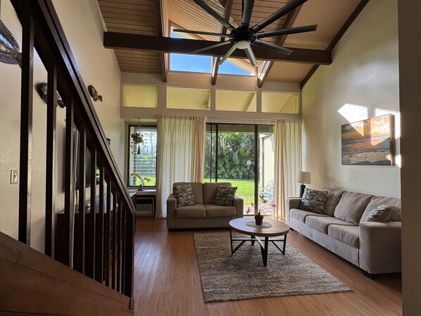 A spacious living room with a high ceiling, a ceiling fan, a large window, and a view of the outdoors.