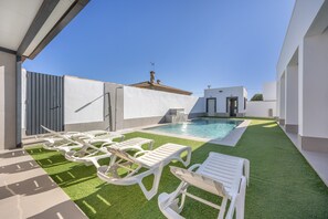 Un espace piscine extérieur moderne, doté d’une pelouse artificielle, de chaises longues blanches et surplombé d’un ciel d’un bleu limpide.