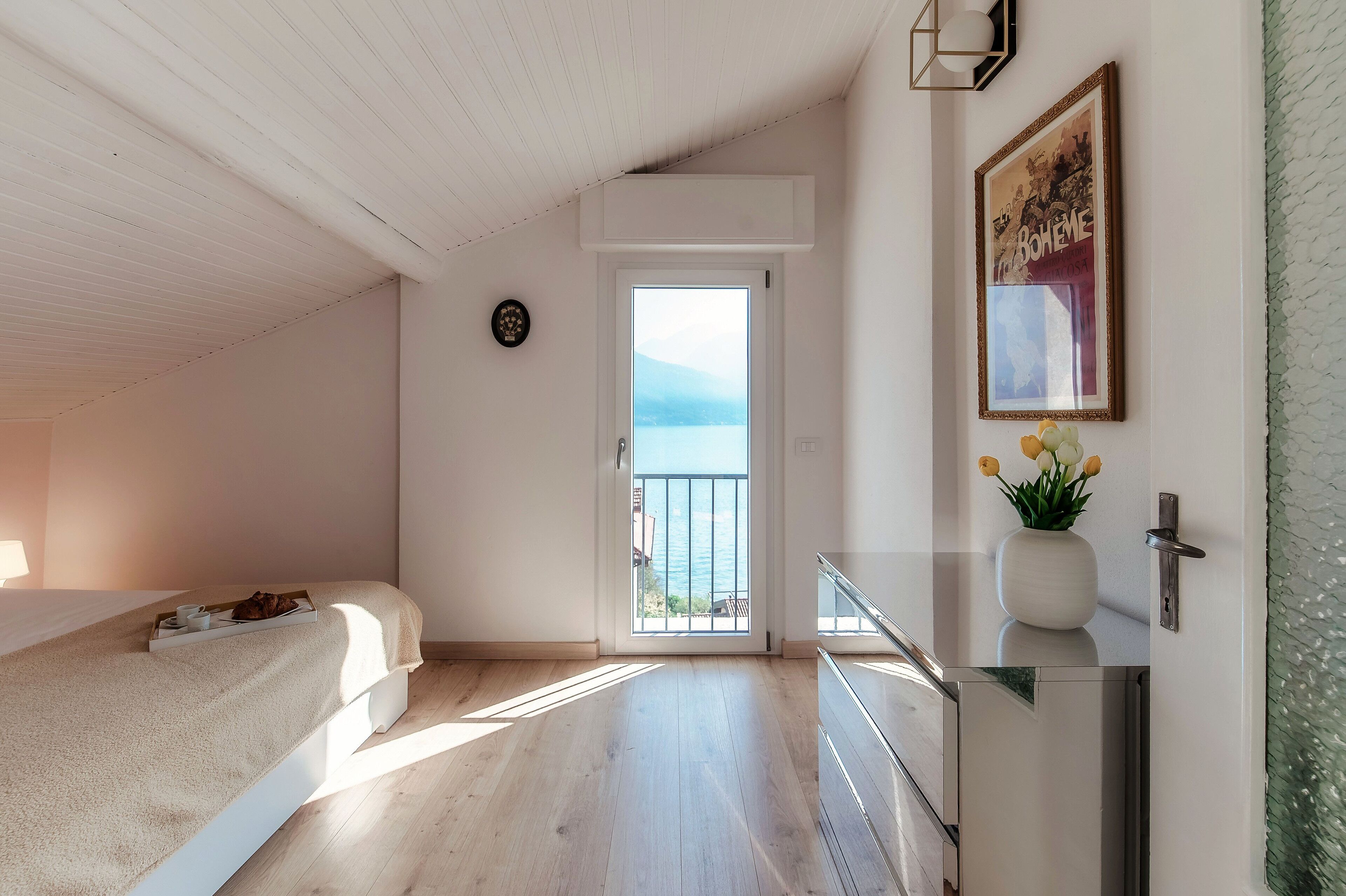 Ein Schlafzimmer mit einem Bett, einem Nachttisch mit einer Vase, einer Tür, die auf einen Balkon führt, und einem gerahmten Bild an der Wand.