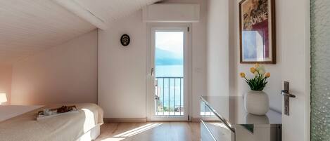 Ein Schlafzimmer mit einem Bett, einem Nachttisch mit einer Vase, einer Tür, die auf einen Balkon führt, und einem gerahmten Bild an der Wand.