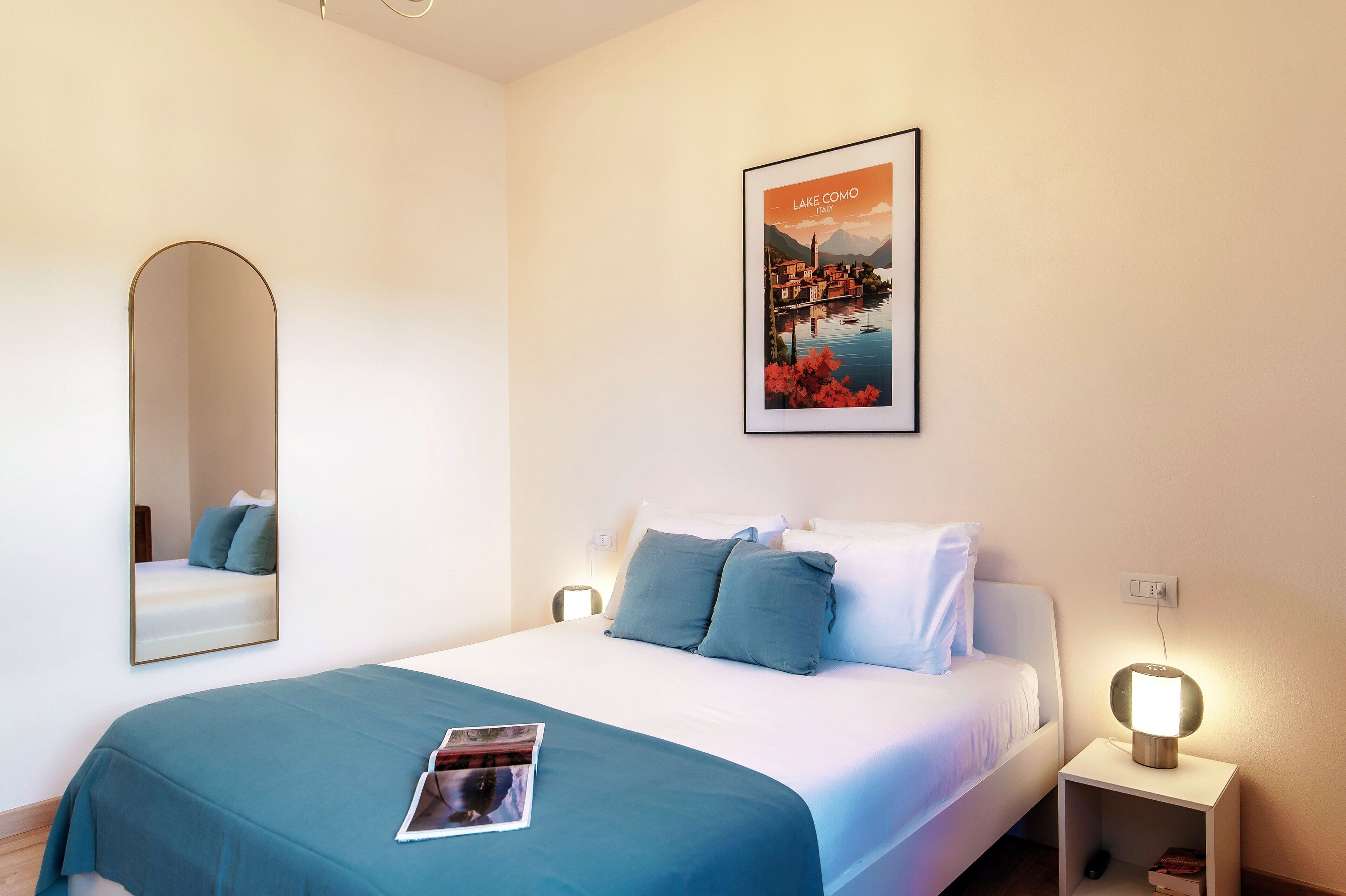 Una cama bien hecha con una colcha y almohadas color aguamarina, un cuadro con una escena de un lago, una lámpara de noche y un espejo en la pared.