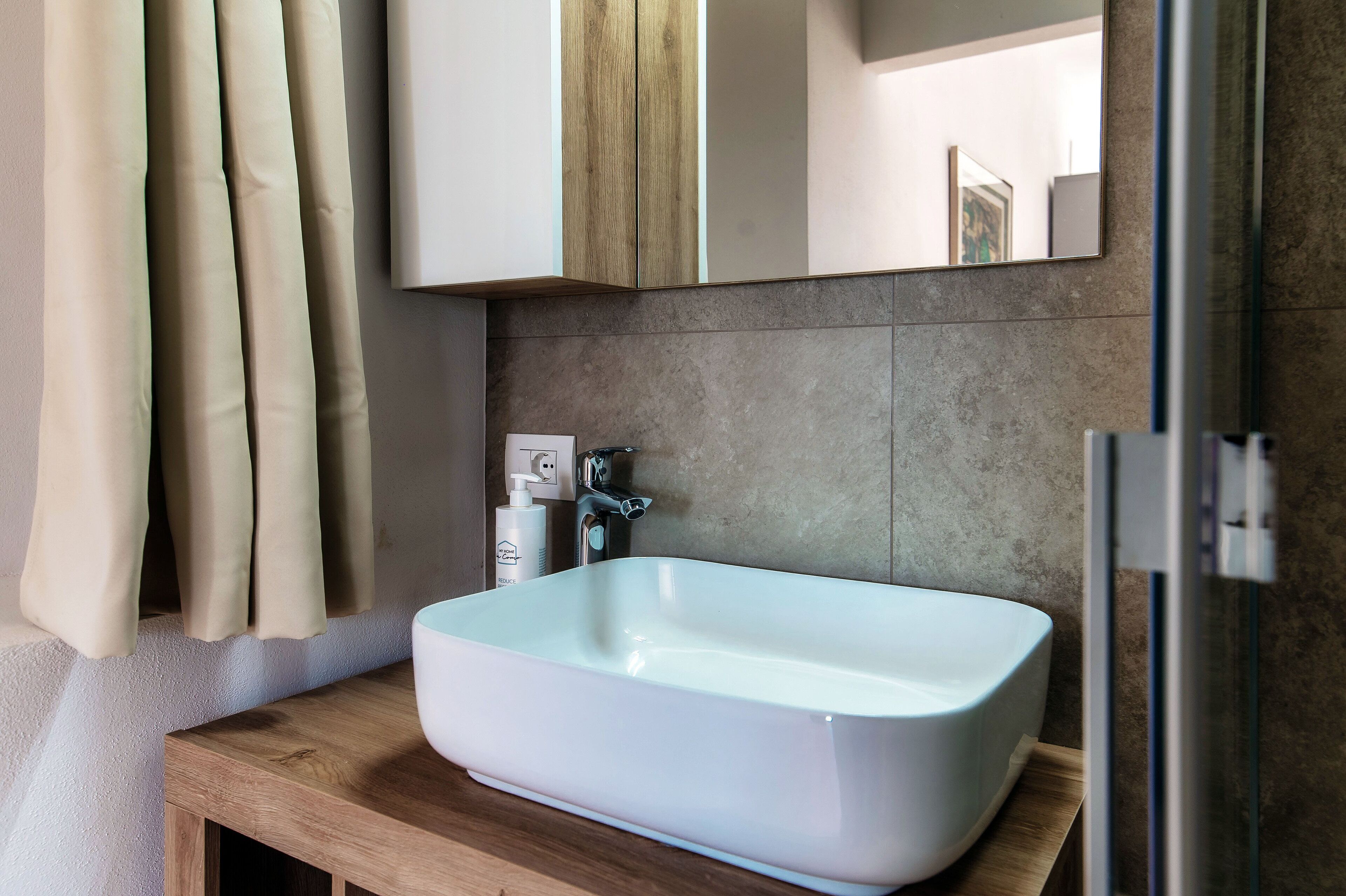 A bathroom with a white sink, wooden countertop, beige curtains, a mirror, and a shower area.