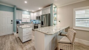 A kitchen with white cabinets, a large island, stainless steel appliances, and a wicker chair. - Sand Pointe 137: Luxury Beachfront Escape with Stunning Gulf Views! (Sanibel)