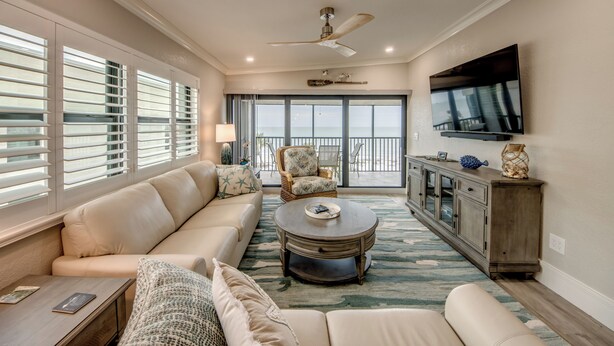 A spacious living room with a large sofa, a coffee table, and a TV mounted on the wall. There is a ceiling fan, a window with blinds, and a view of the outdoors. - Sand Pointe 137: Luxury Beachfront Escape with Stunning Gulf Views! (Sanibel)