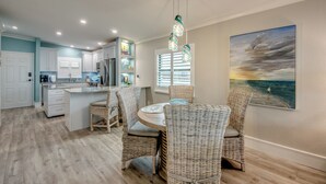 A modern kitchen with white cabinets, a dining area with wicker chairs, and a large painting of a beach scene. - Sand Pointe 137: Luxury Beachfront Escape with Stunning Gulf Views! (Sanibel)