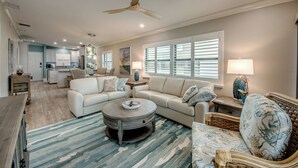 A spacious living area with a large round coffee table, a sofa, and a wicker chair. There is a kitchen in the background with white cabinets and a dining area. - Sand Pointe 137: Luxury Beachfront Escape with Stunning Gulf Views! (Sanibel)